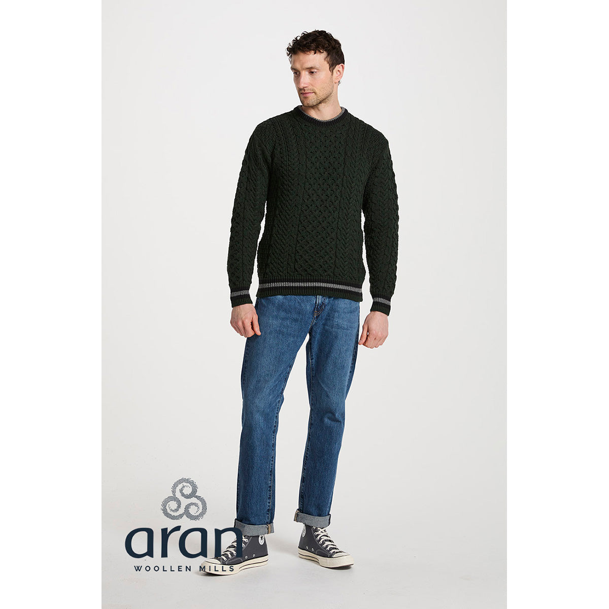 Man modeling a dark green Merino Aran crewneck sweater with cable knit pattern and striped hem and cuffs.
