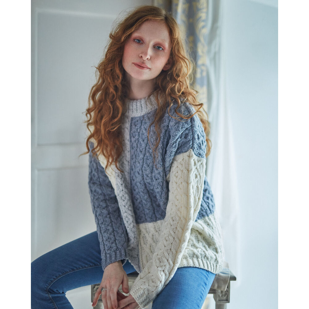 Woman wearing a chunky three color patchwork cable knit sweater in blue gray and cream.