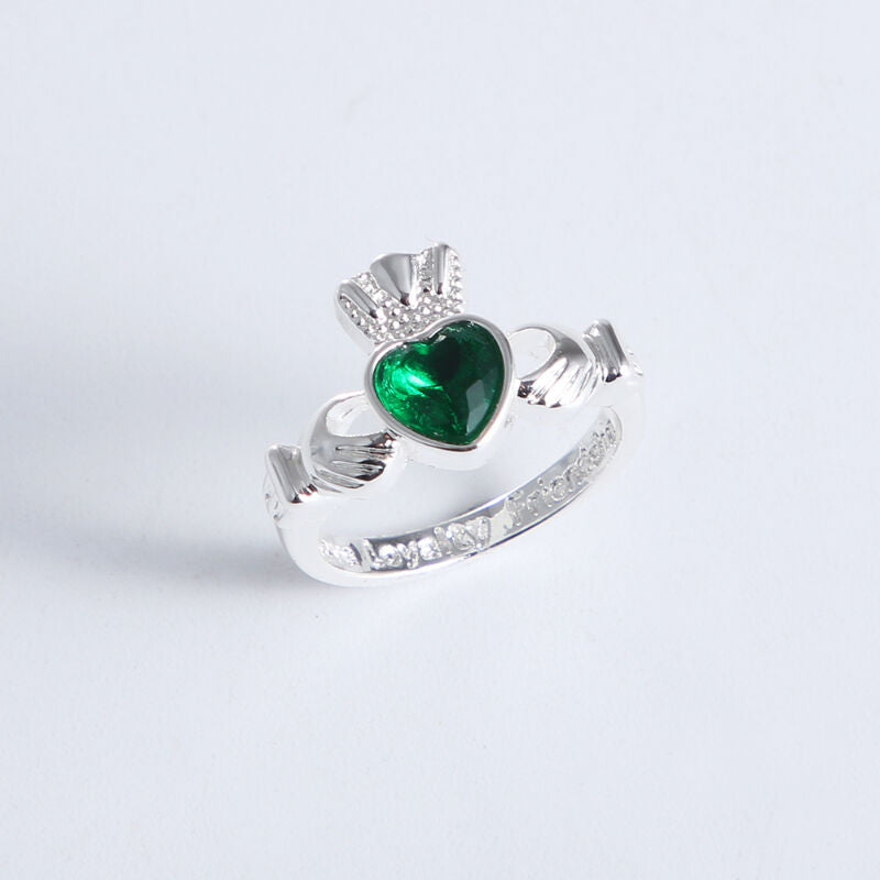 Silver ring with green heart-shaped gem and crown held by two hands