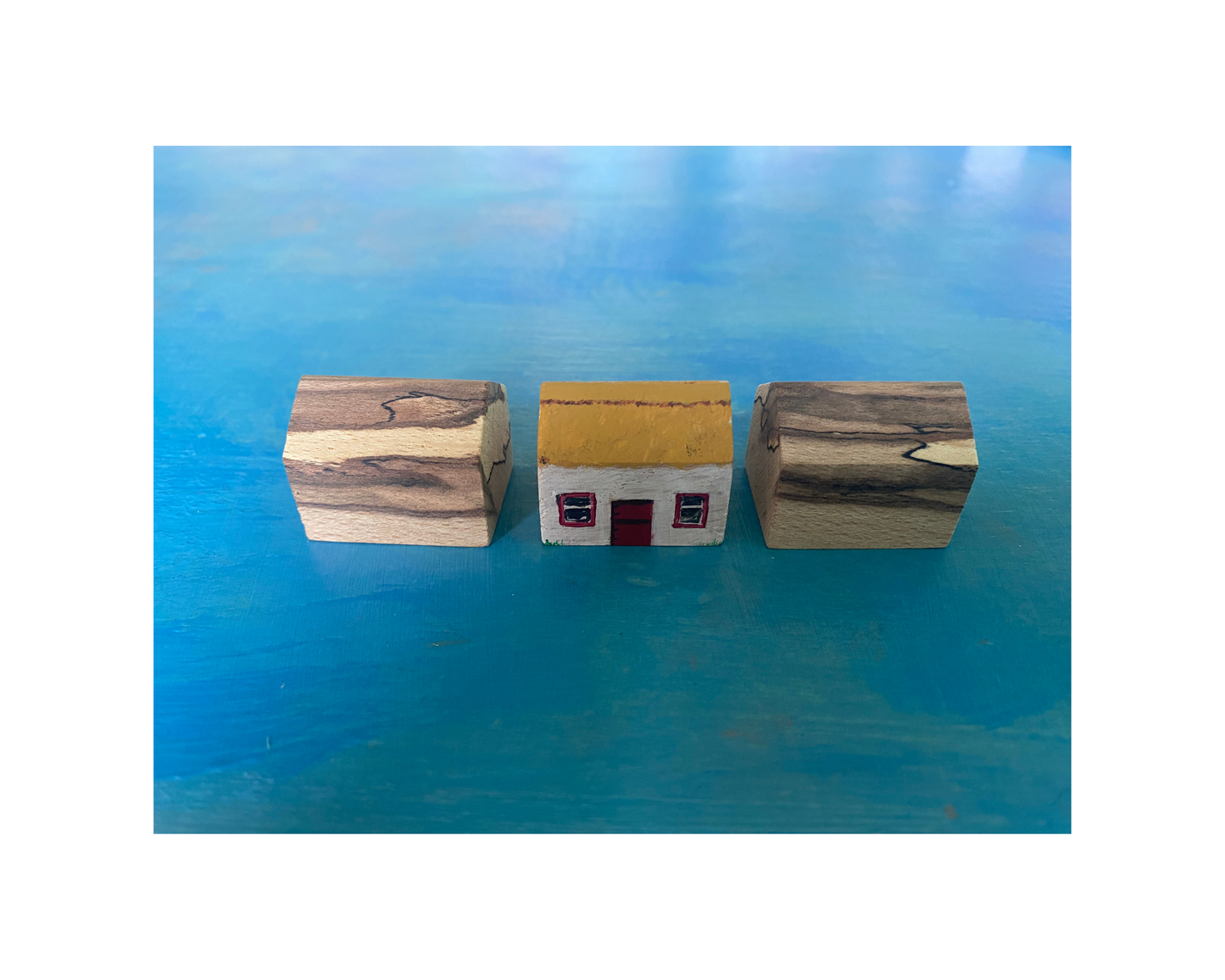Three wooden cottage magnets on a blue surface; middle magnet shows a small house with a yellow roof and red door.