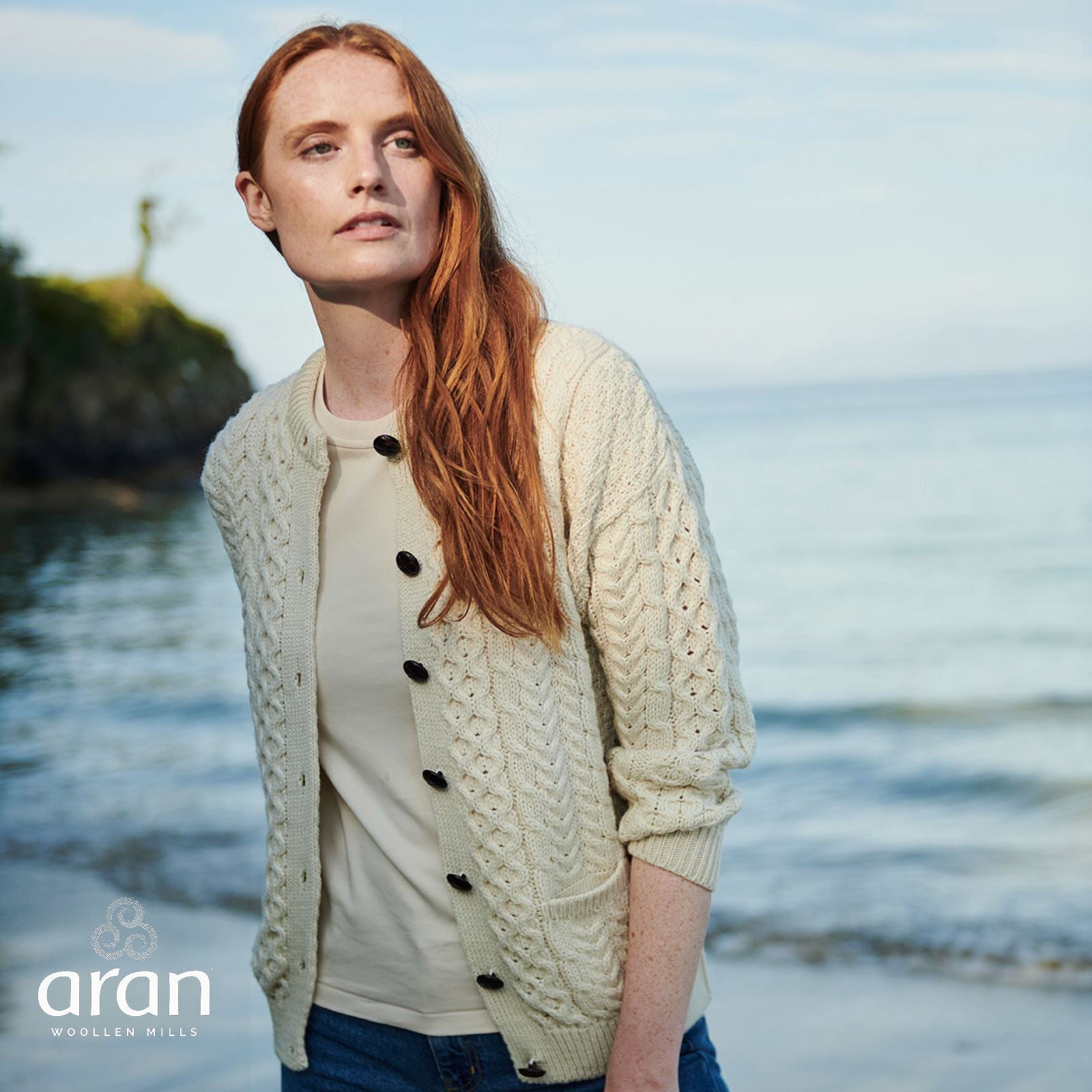 Cream Aran cardigan with pockets worn closed over a light top by the sea.