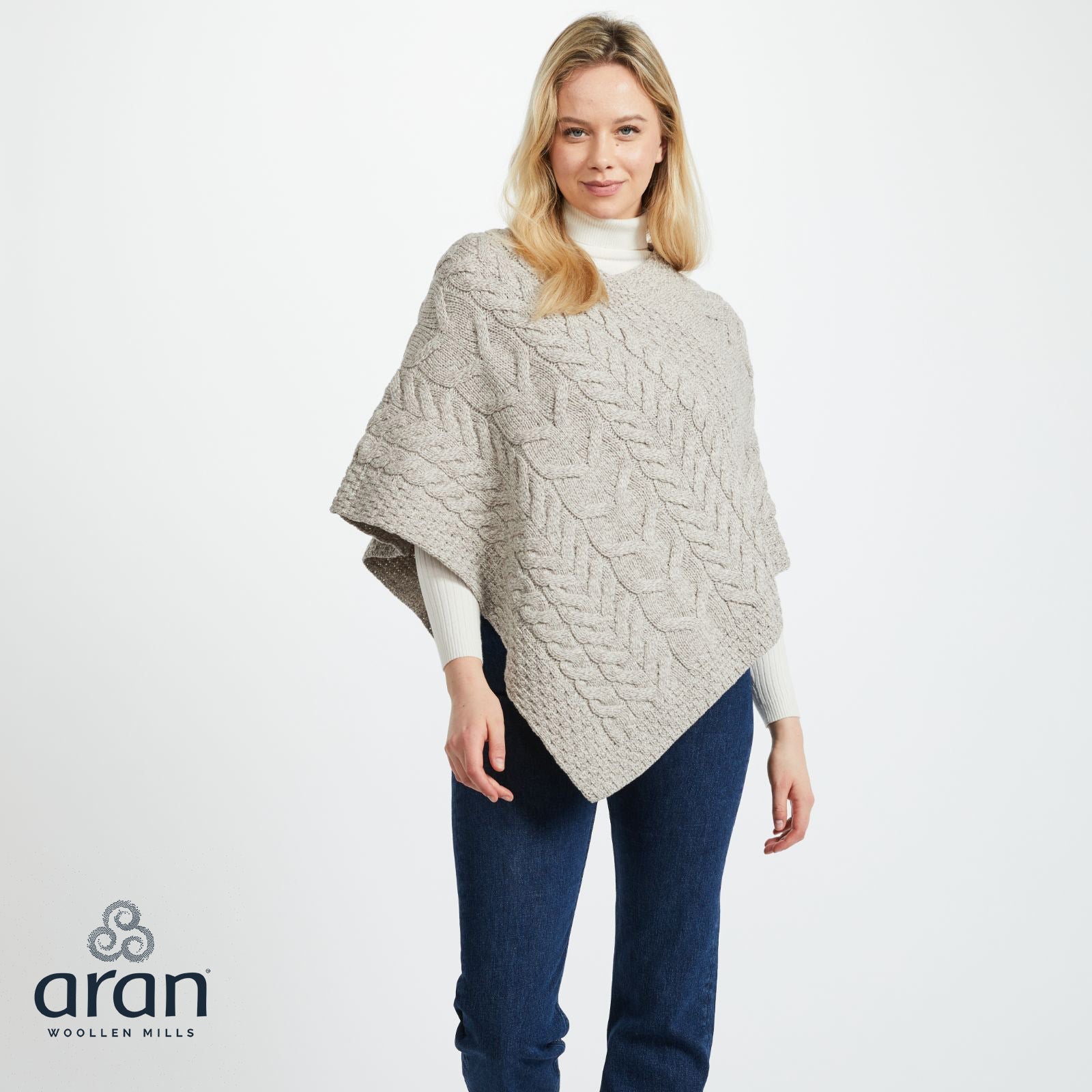 Three-quarter view of a model wearing an oatmeal Aran poncho over a white ribbed sweater and blue jeans.