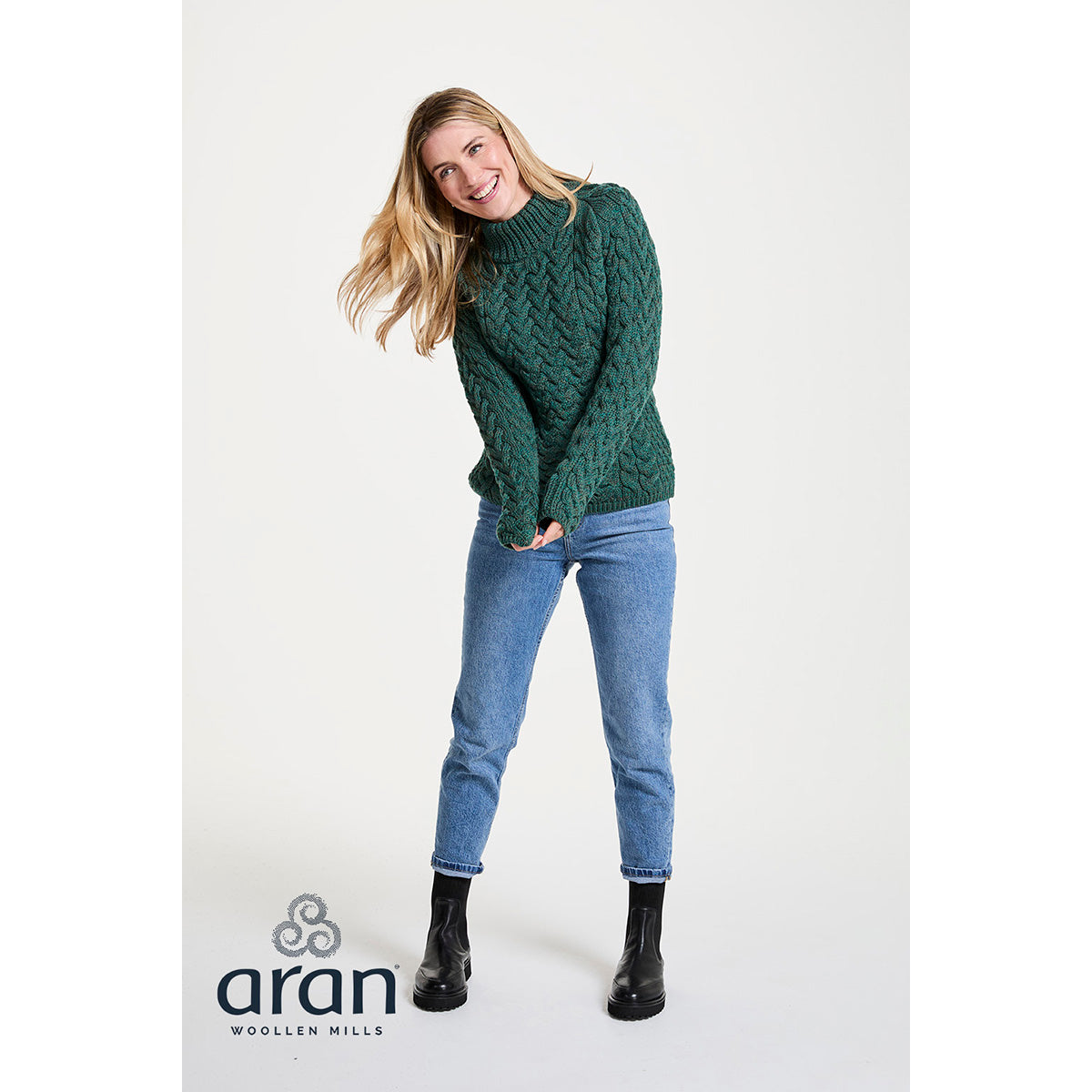 Full body shot of a woman in a green Knightstown Merino Aran Crew sweater with jeans and black boots