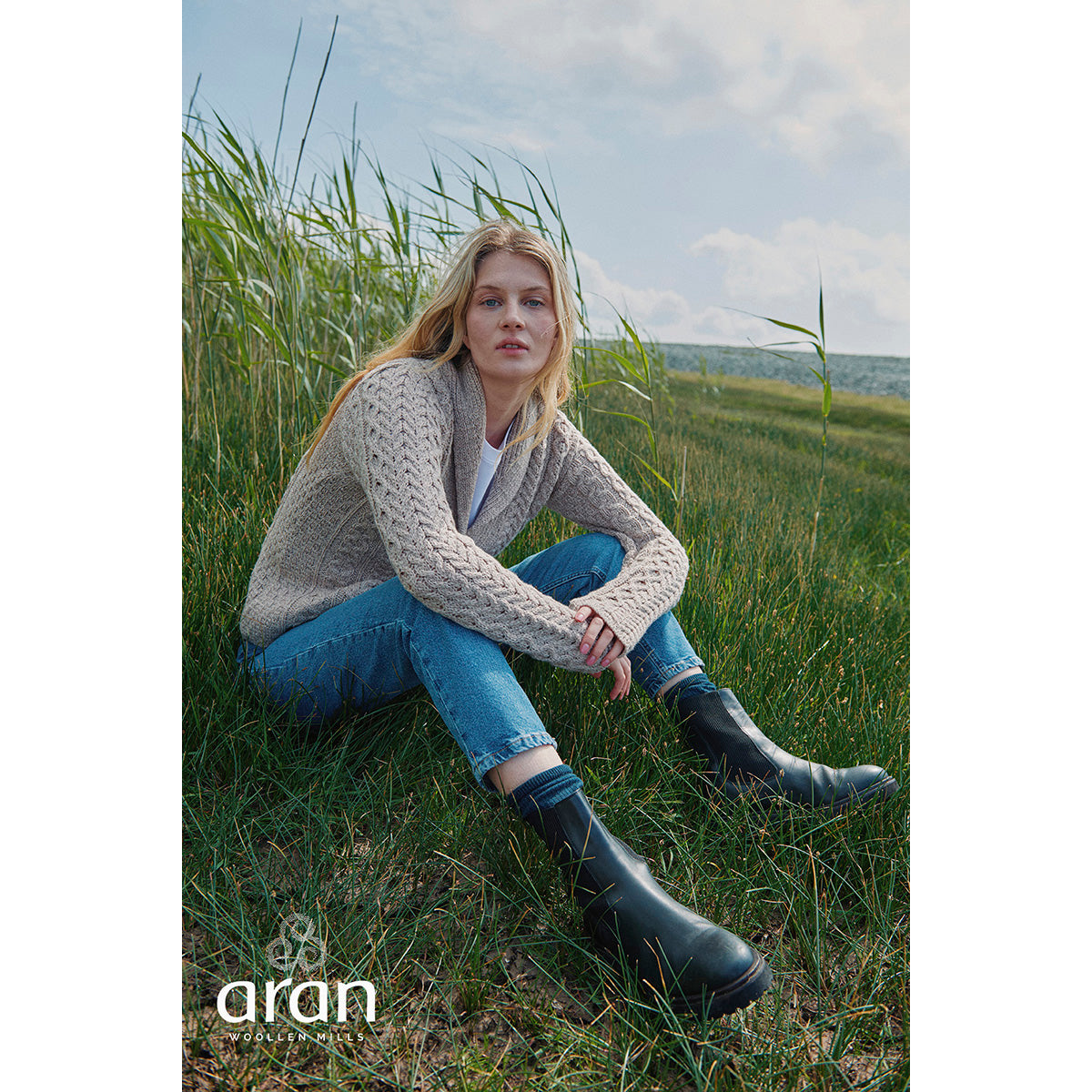 Woman wearing a beige shawl collar Merino Aran cardigan sits in tall coastal grasses wearing blue jeans and black knee high boots.