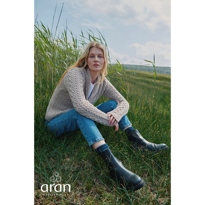 Woman wearing a beige shawl collar Merino Aran cardigan sits in tall coastal grasses wearing blue jeans and black knee high boots.