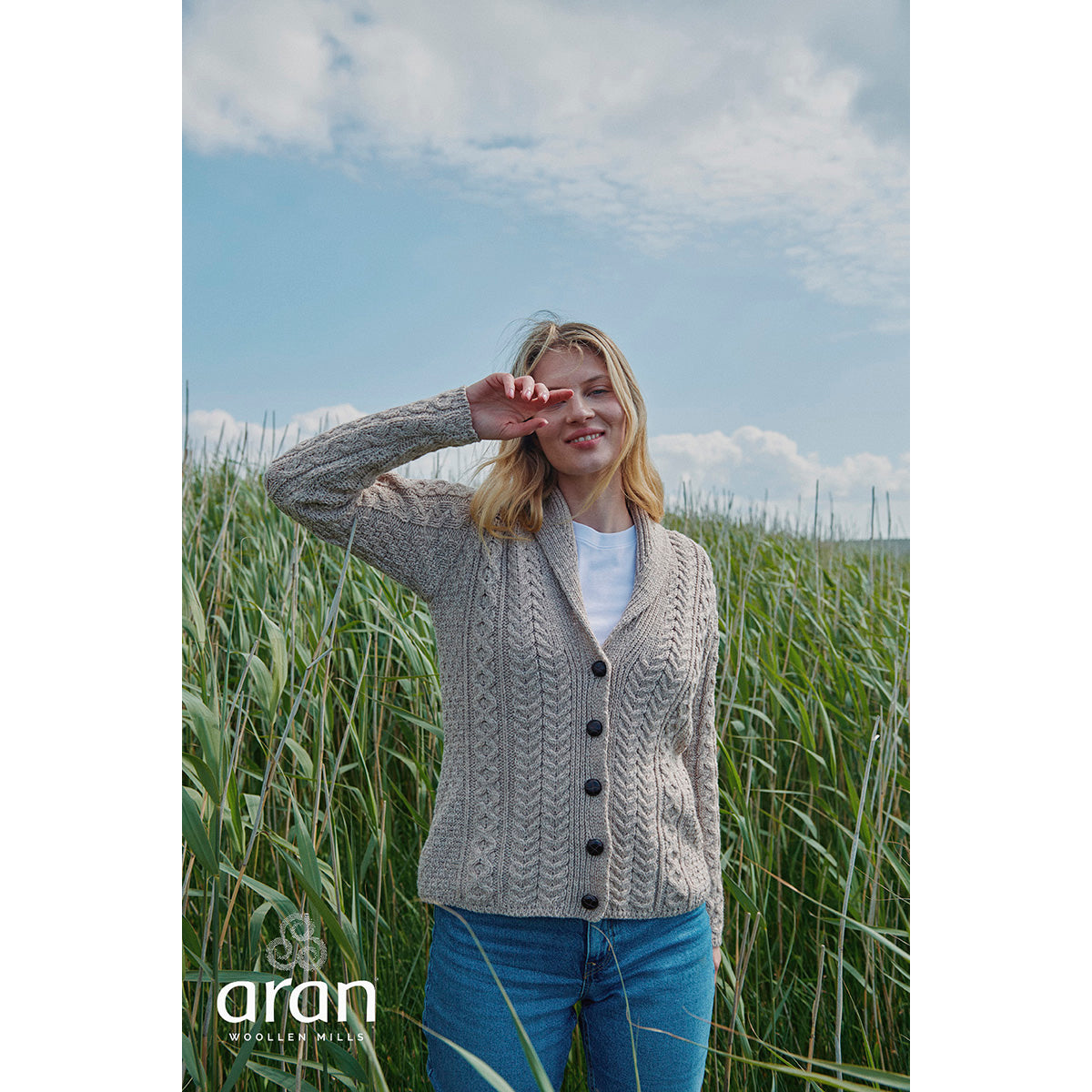 Womens Tourmakeady Shawl Collar Merino Aran Cardigan in beige knit worn by a model in a grassy field.
