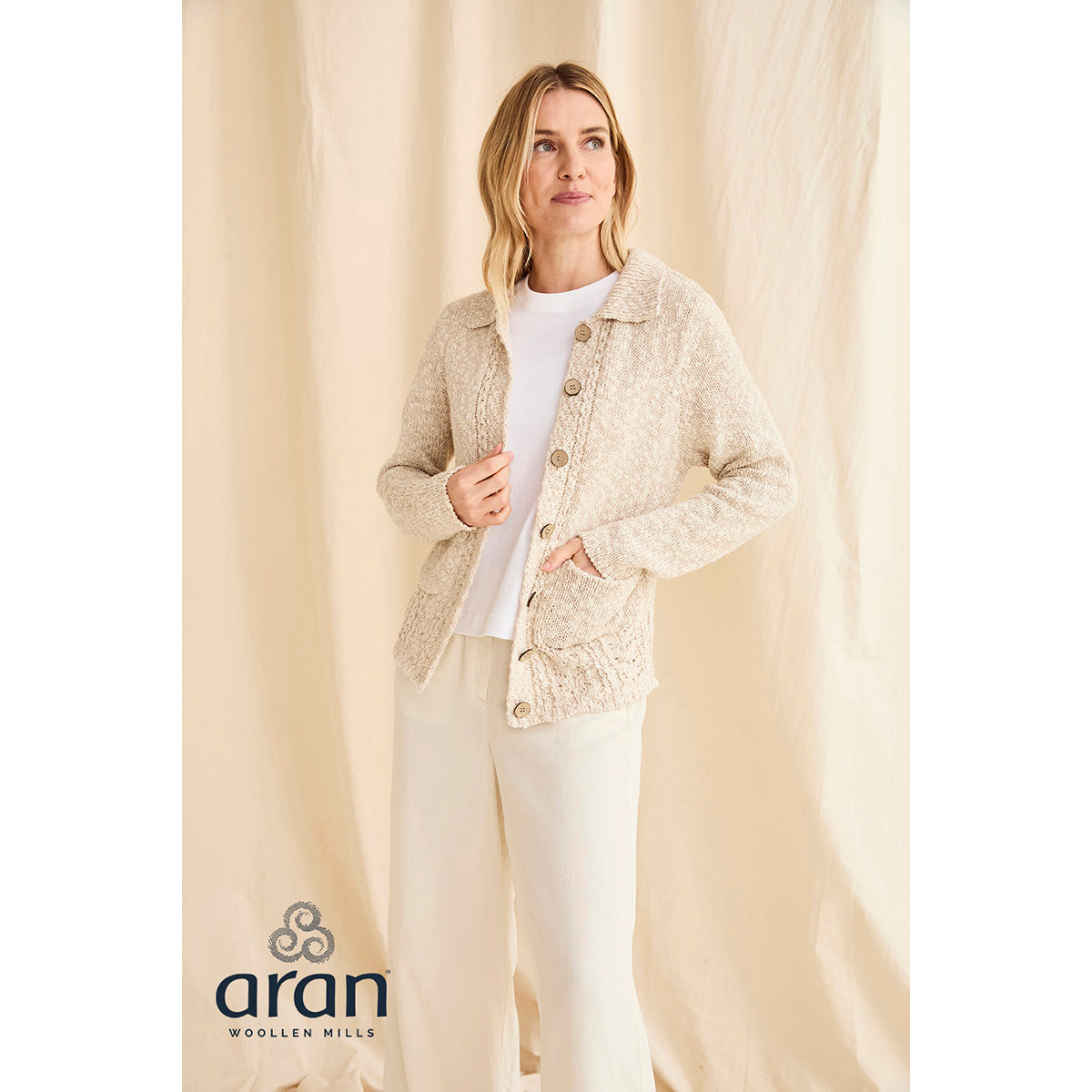 Full length view of a woman wearing the cream Eriu cardigan with pockets and buttons against a beige backdrop.