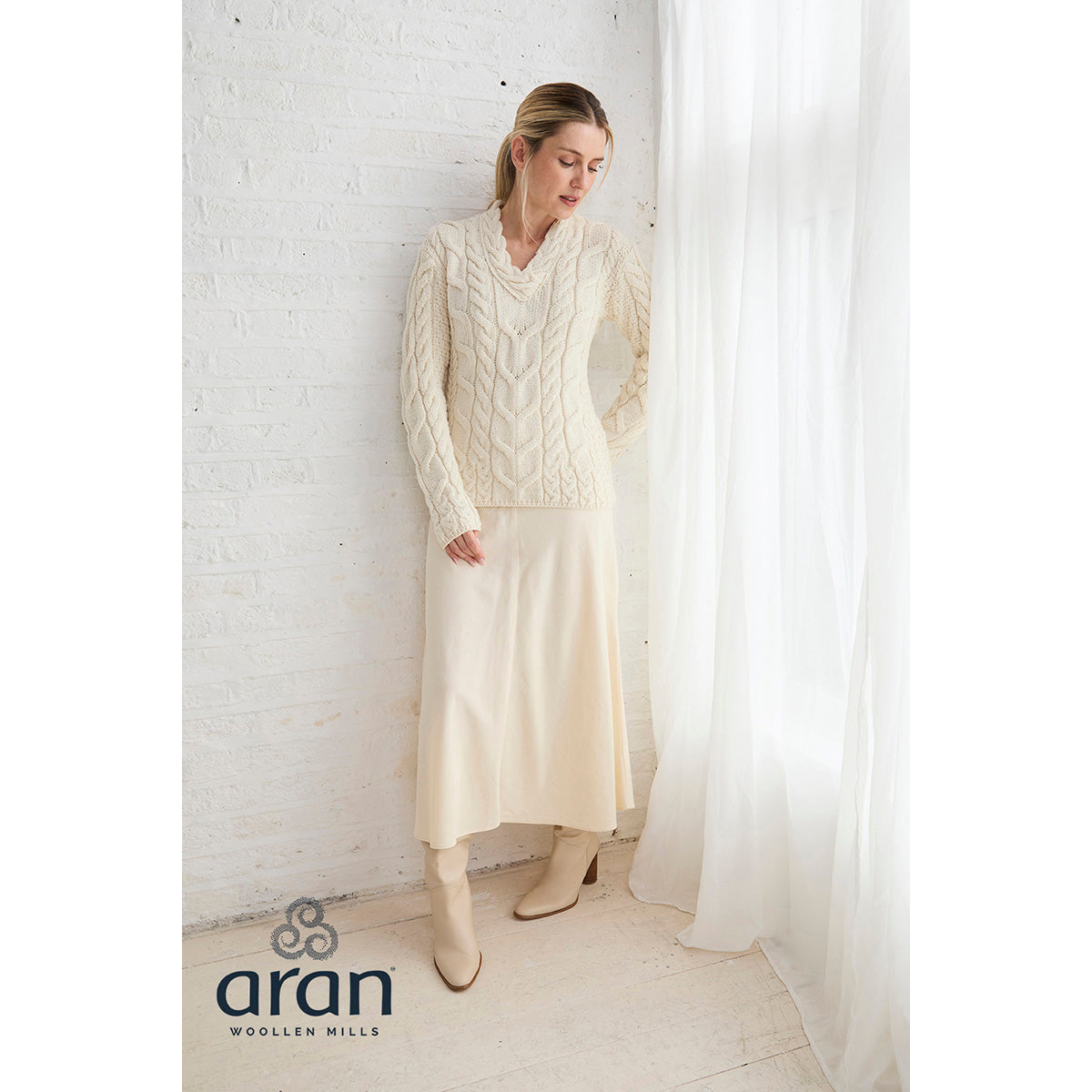 Full-length view of model in cream Tralee sweater with a matching skirt and boots beside a white wall and sheer curtain Aran Woollen Mills logo visible