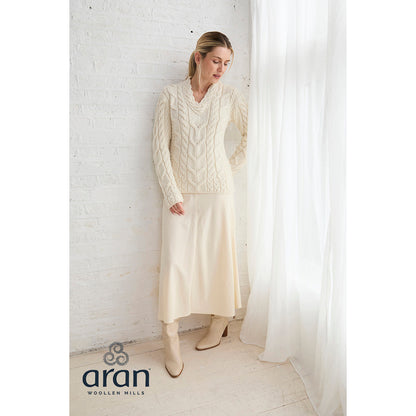 Full-length view of model in cream Tralee sweater with a matching skirt and boots beside a white wall and sheer curtain Aran Woollen Mills logo visible