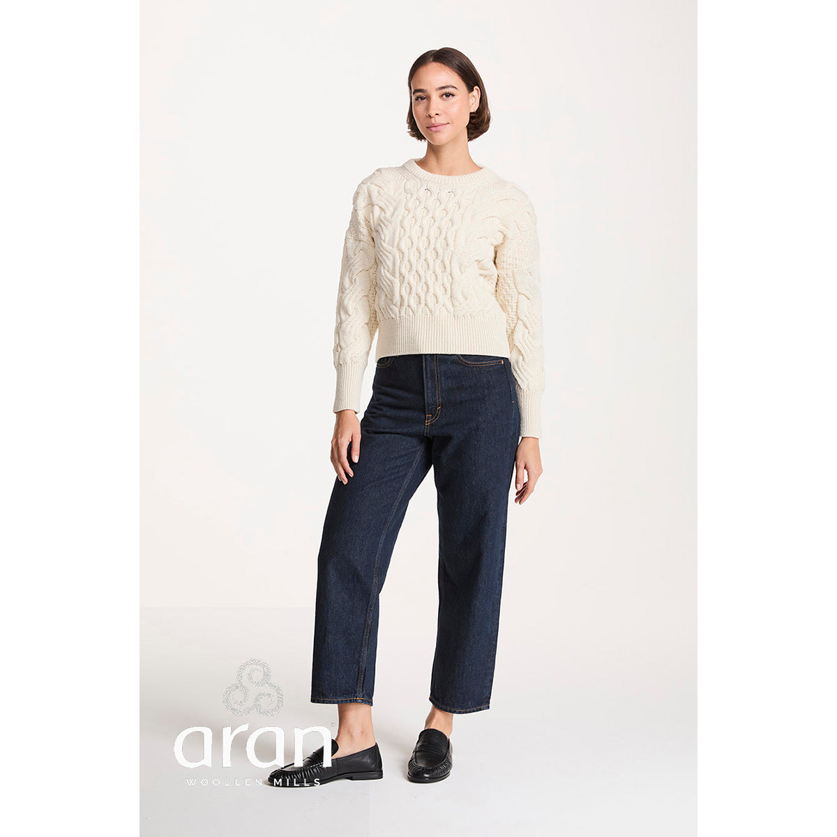 Full body product shot of the cream cropped cable knit sweater on a model with dark denim jeans and black loafers.