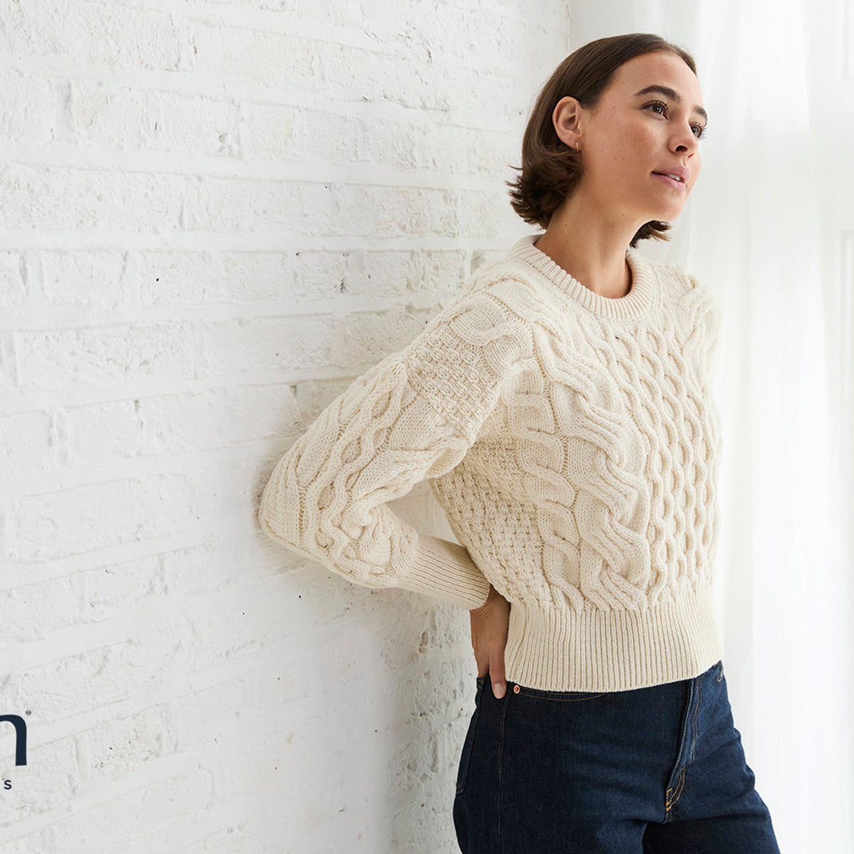 Female model in cream cropped cable knit sweater standing against a white brick wall.