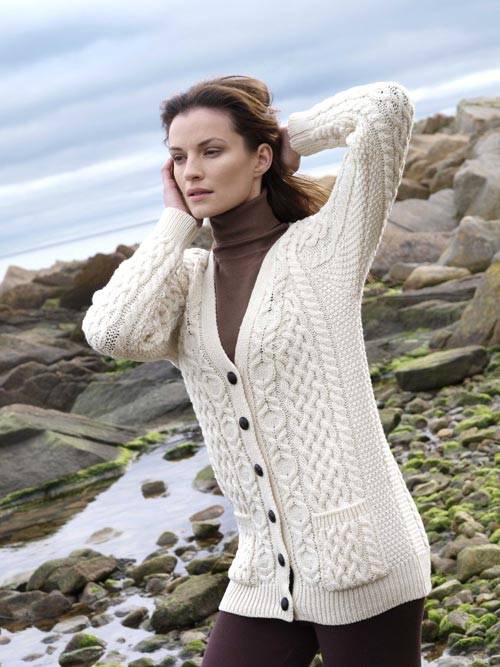 Model wearing a cream Aran cardigan over a brown turtleneck posing on a rocky shoreline.