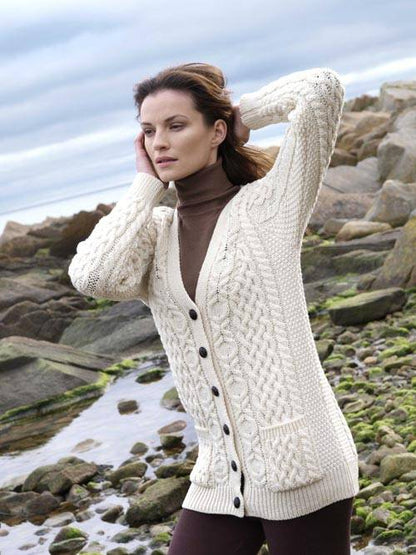 Model wearing a cream Aran cardigan over a brown turtleneck posing on a rocky shoreline.