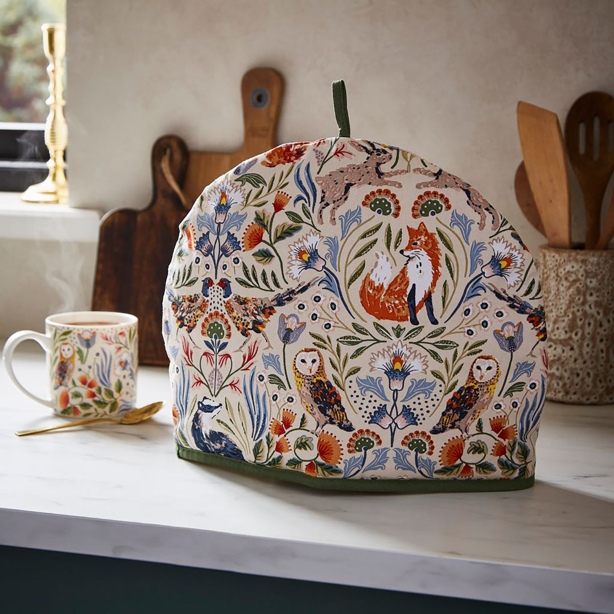 Blackthorn Tea Cozy with a woodland pattern featuring foxes, owls, and flowers on cream fabric with a green trim.