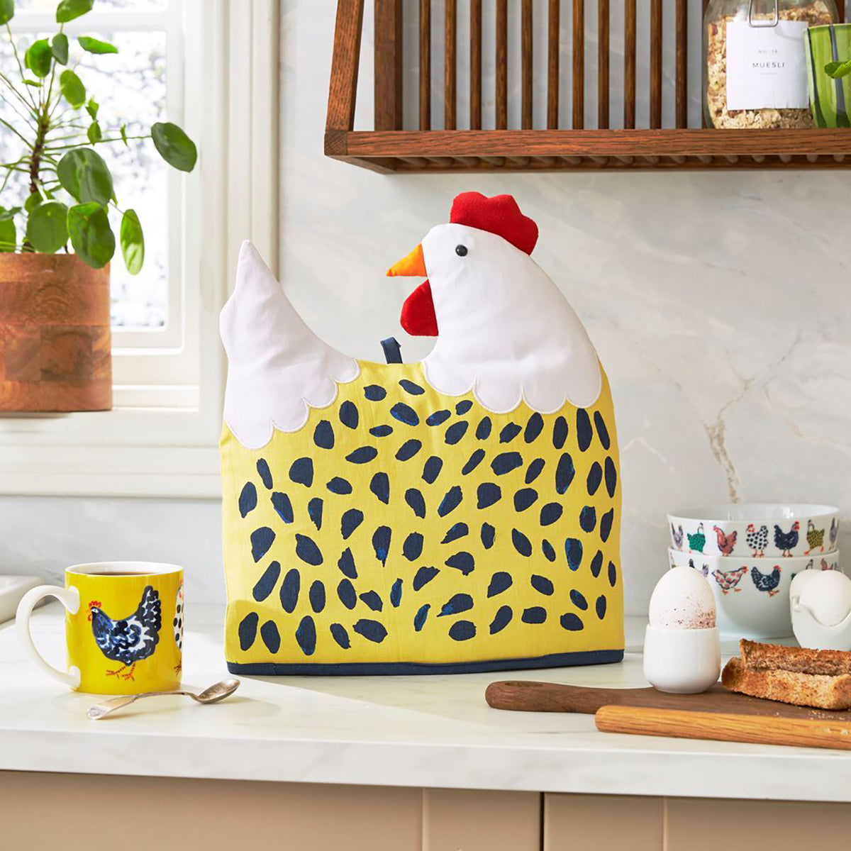 Chick Chicken Shaped Tea Cozy in yellow stands on a kitchen counter beside eggs and bowls.