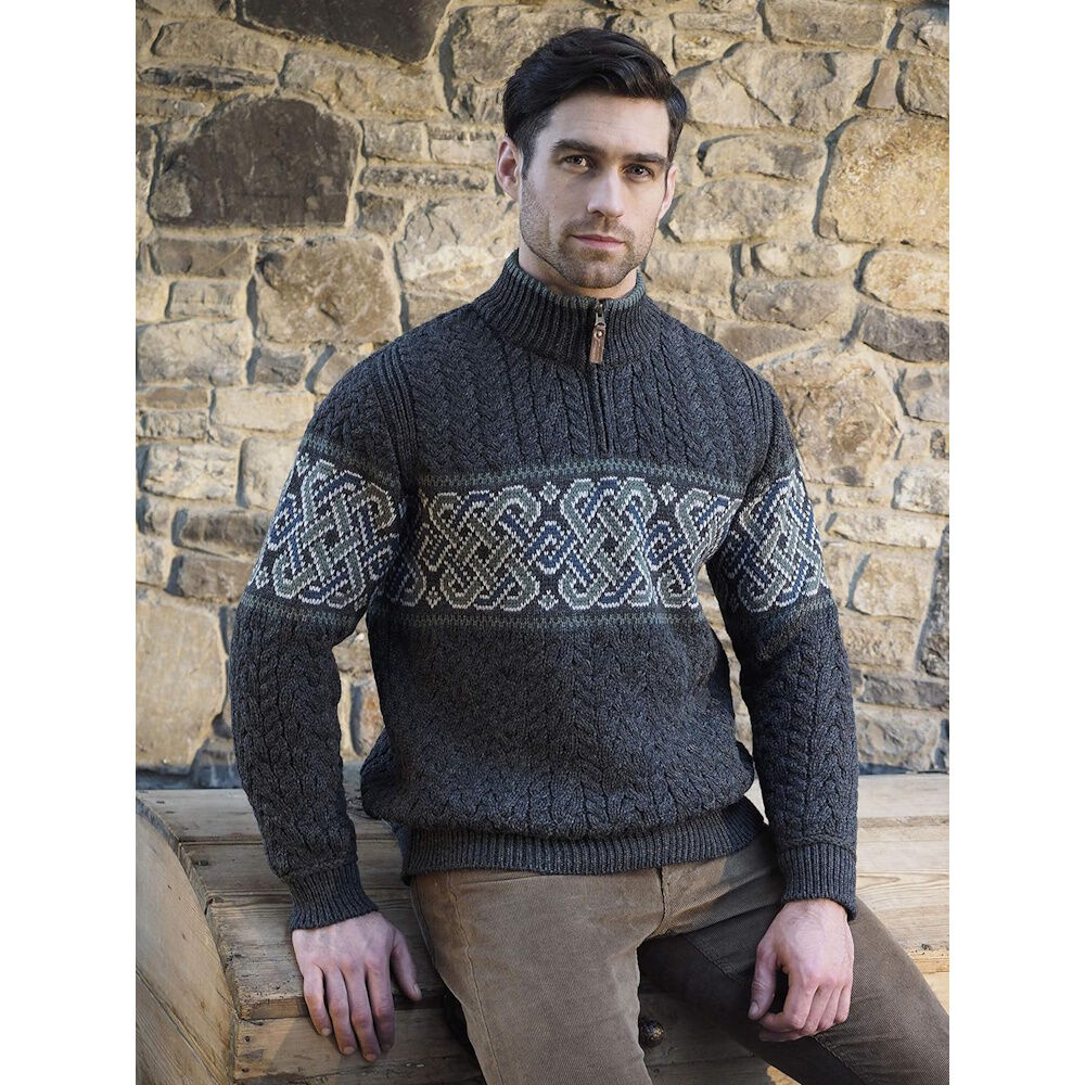 Man in charcoal Aran sweater with a bold Celtic band across the chest leaning against a stone wall