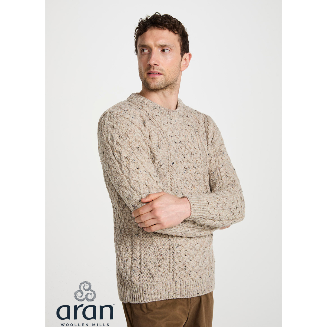 Male model in beige oatmeal fleck Aran crewneck sweater seen in side profile with arms crossed.