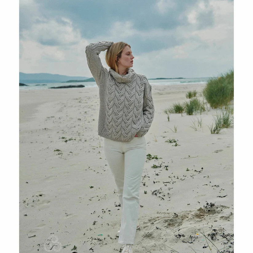 Woman on a beach wearing an oatmeal chunky cable Aran sweater.