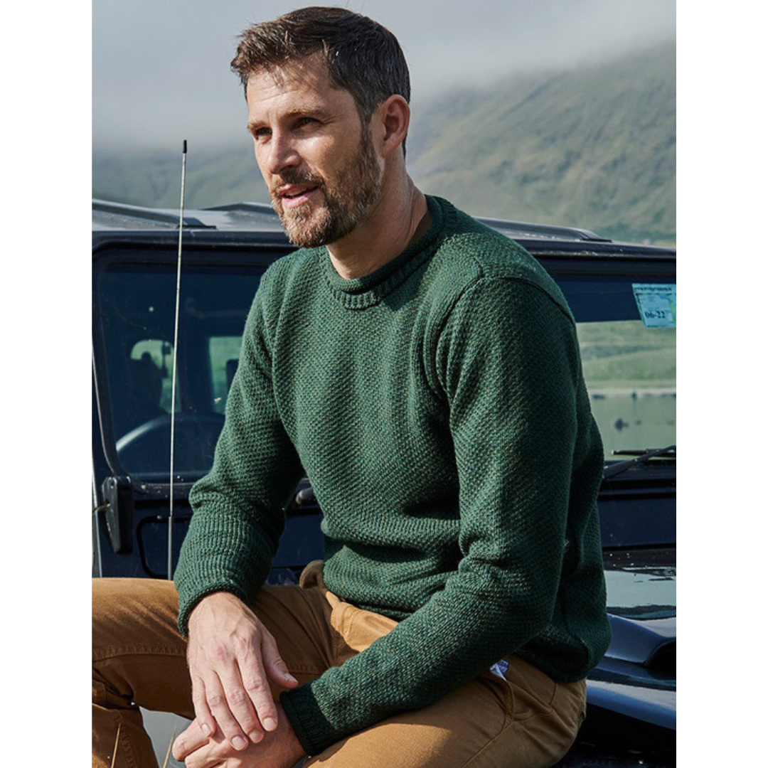 Close up of a man in a green merino wool roll neck sweater leaning on a jeep against rugged green hills.