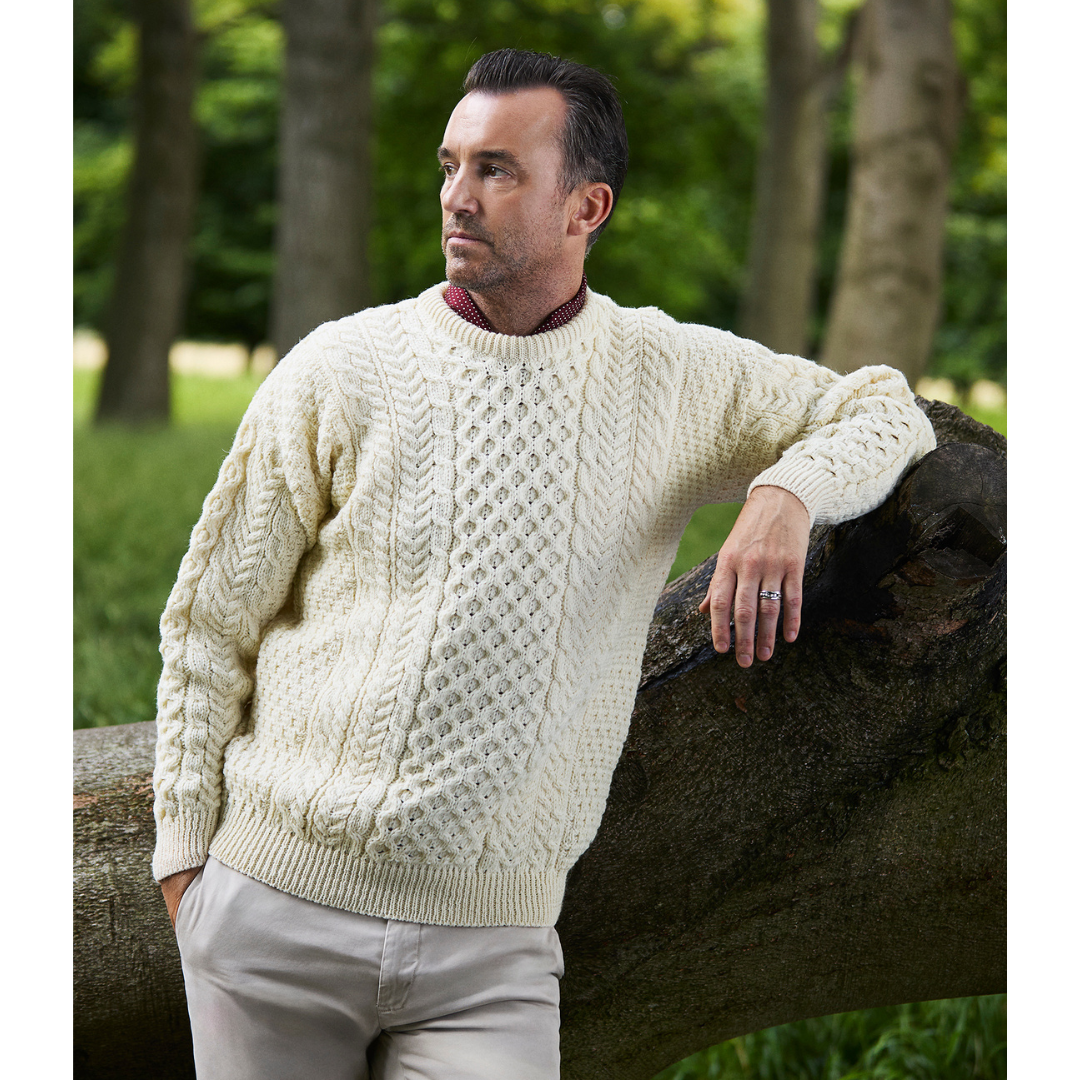 Man leaning on a tree trunk wearing a cream natural chunky cable knit crew neck sweater outdoors.