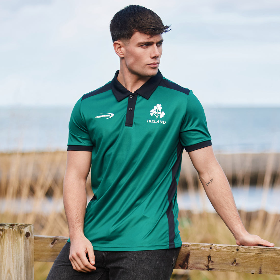 Male model wearing Ireland green and black polo shirt outdoors by a wooden railing