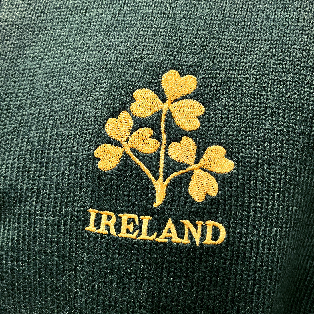 Close up of gold shamrock embroidery on dark green knit with IRELAND text.