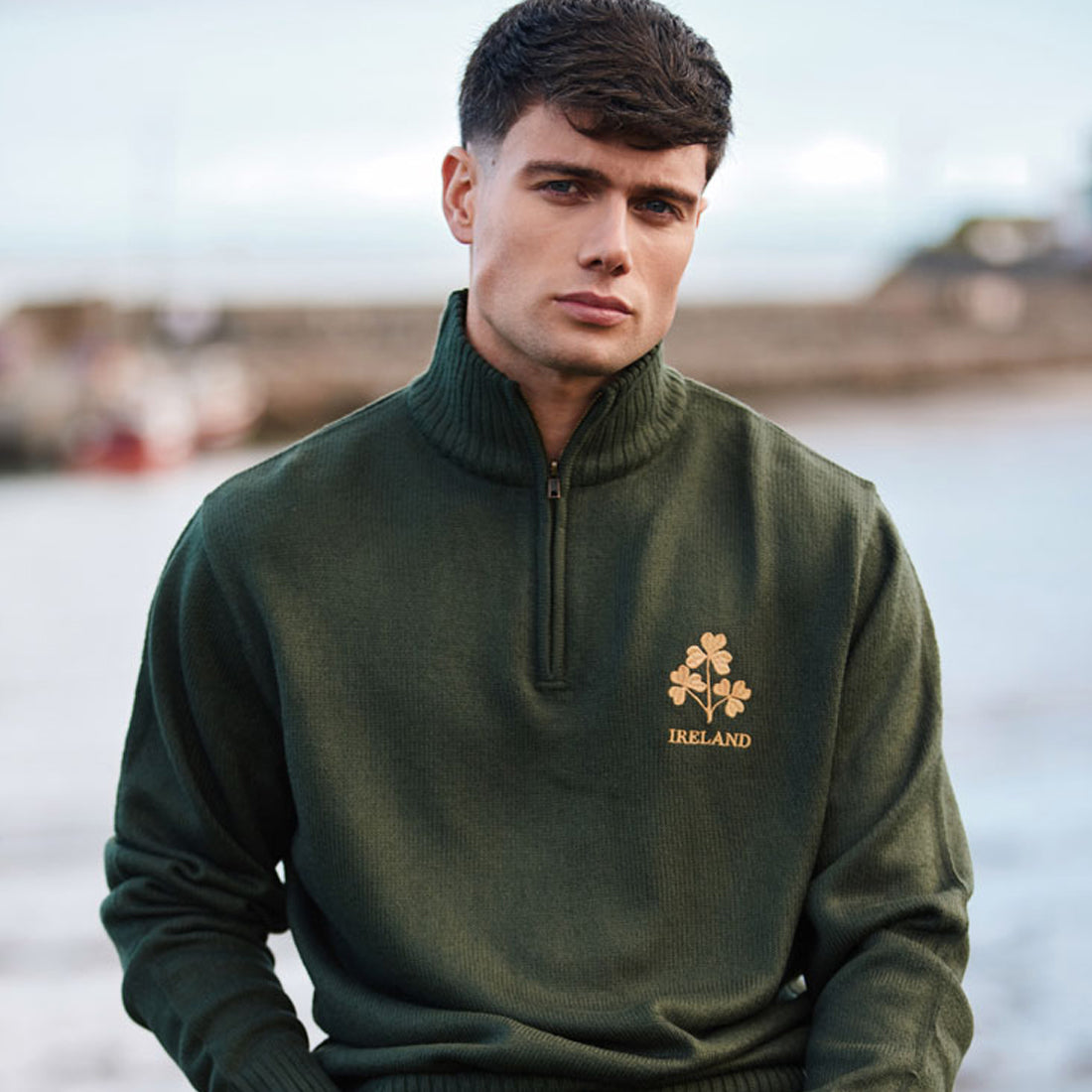 Man modeling a bottle green Ireland jumper with a gold shamrock emblem on the chest by the waterfront.