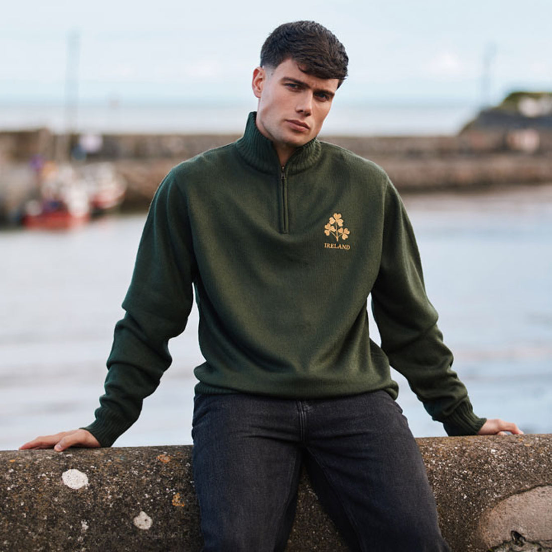 Man sits on a seawall wearing a bottle green quarter zip jumper with gold shamrock Ireland emblem.