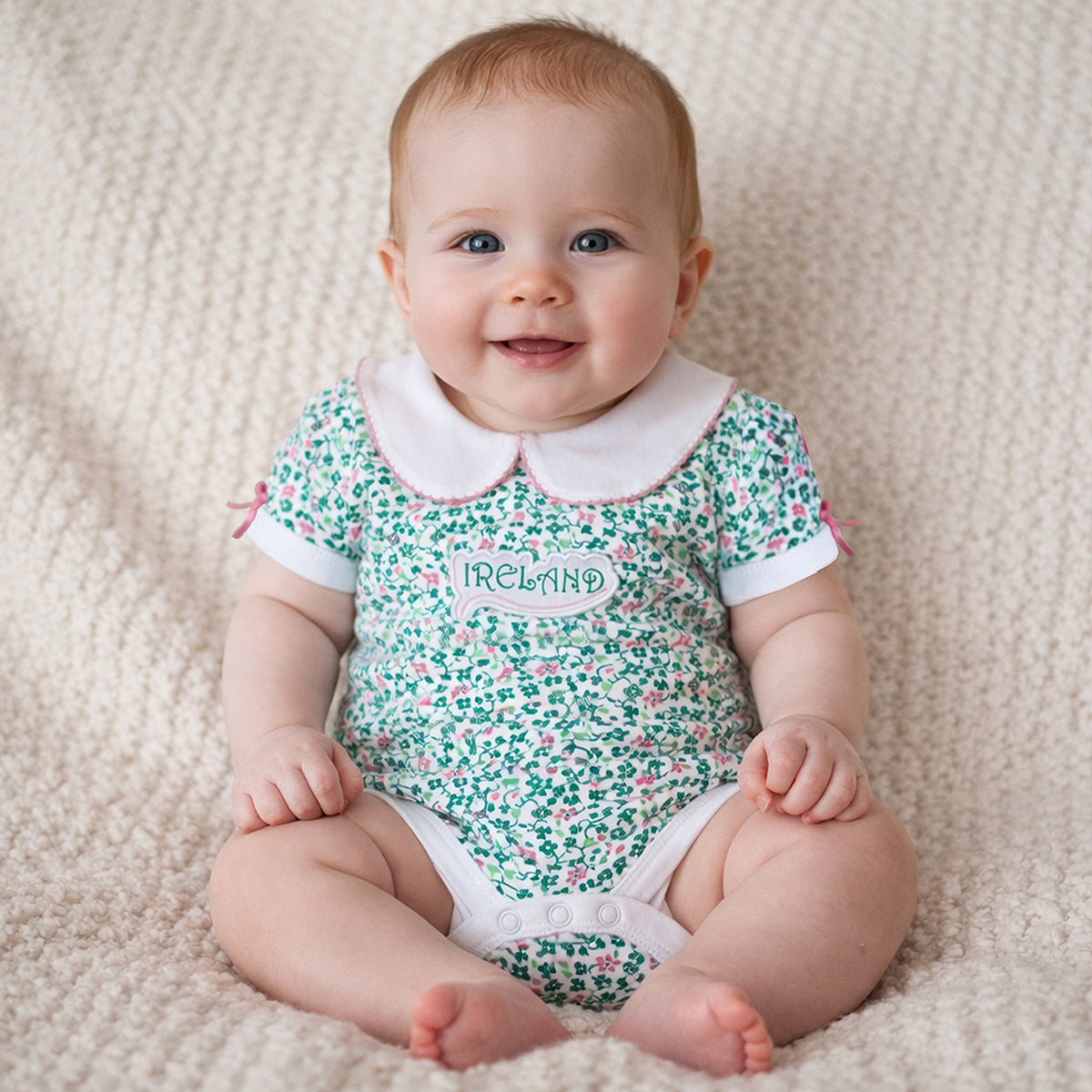 Smiling baby wearing a green floral shamrock print vest with a white collar on a beige rug