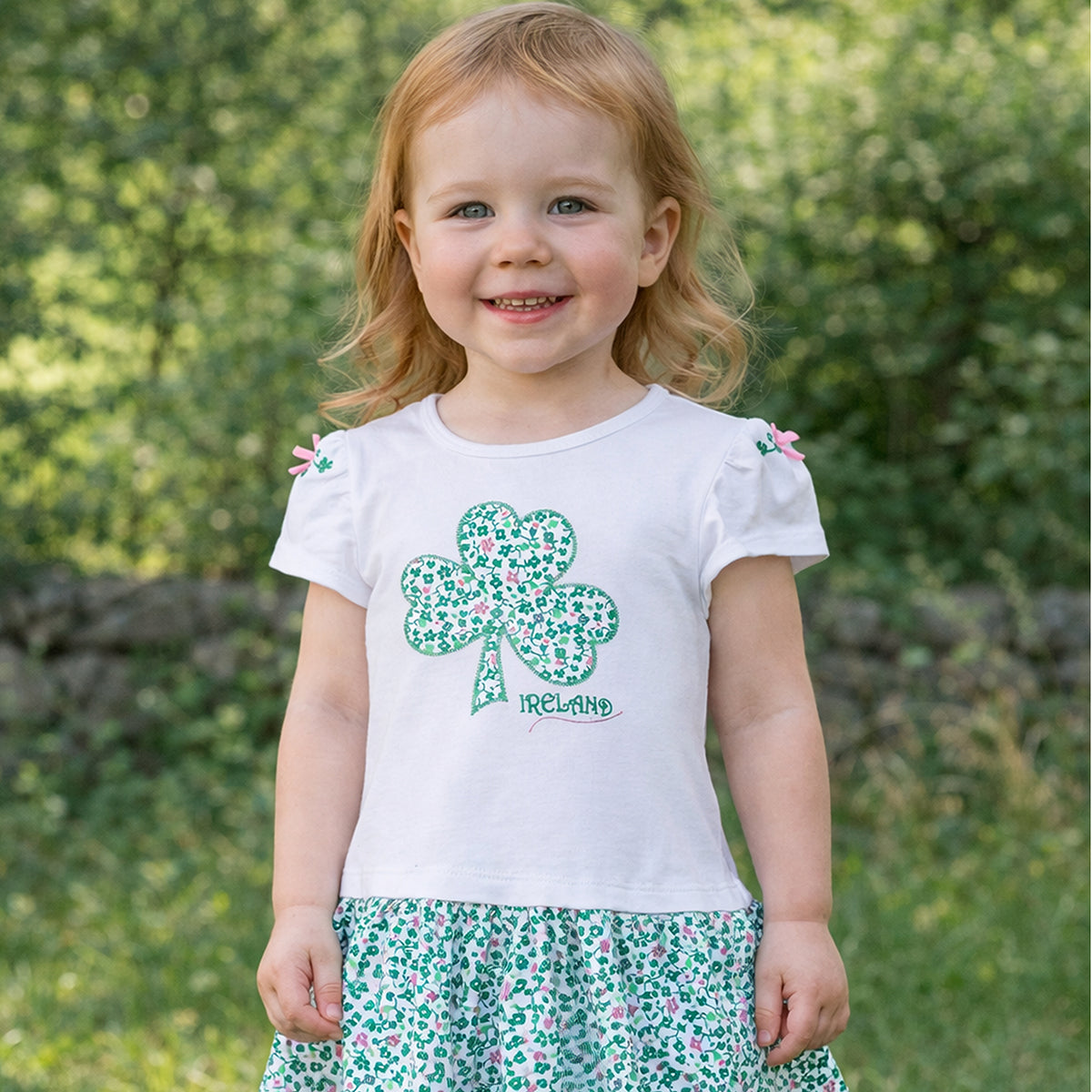 Smiling toddler wearing a white Shamrock Applique Kids Frill Dress with a green floral skirt outdoors.