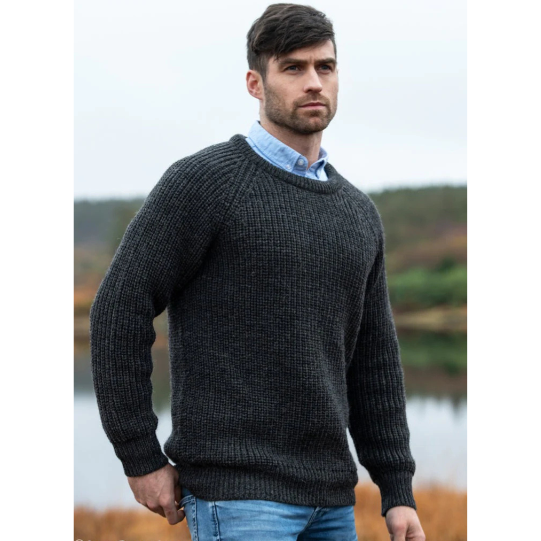 Man in a charcoal fisherman ribbed Aran sweater posing outdoors by a lake.