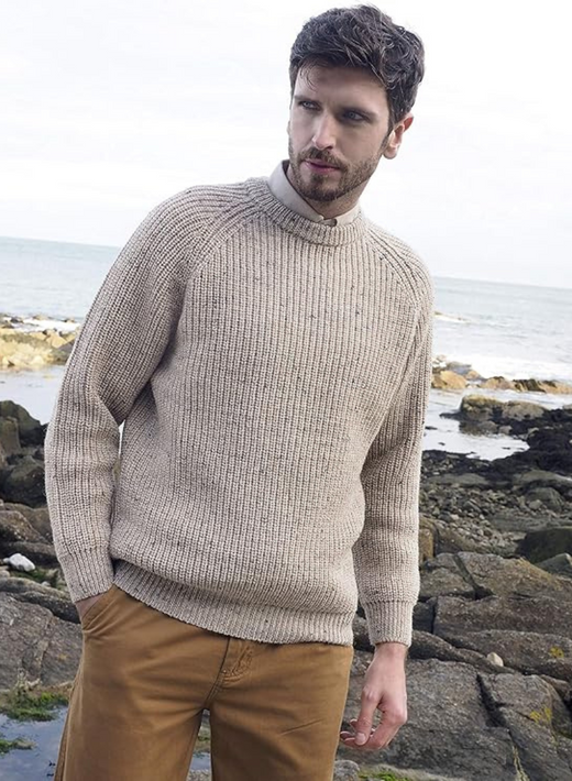 Male model wearing beige fisherman's ribbed Aran sweater standing on a rocky shoreline.