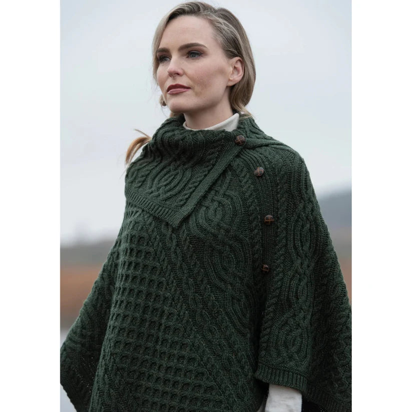 Model wearing a dark green chunky knit poncho with a high cowl neck and side buttons.