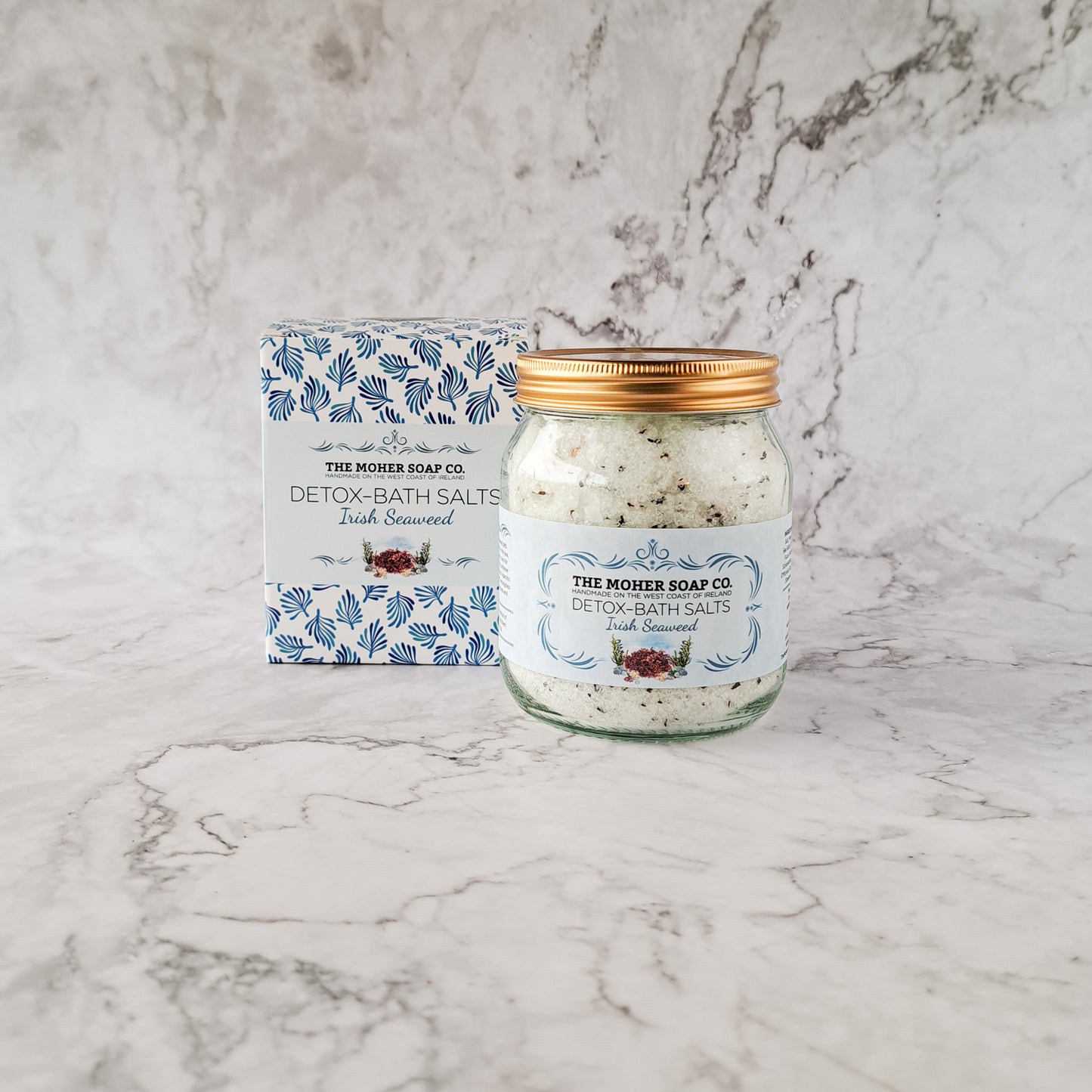 The Moher Soap Co Irish Seaweed Detox Bath Salts jar with a gold lid beside its blue patterned box on a marble surface.