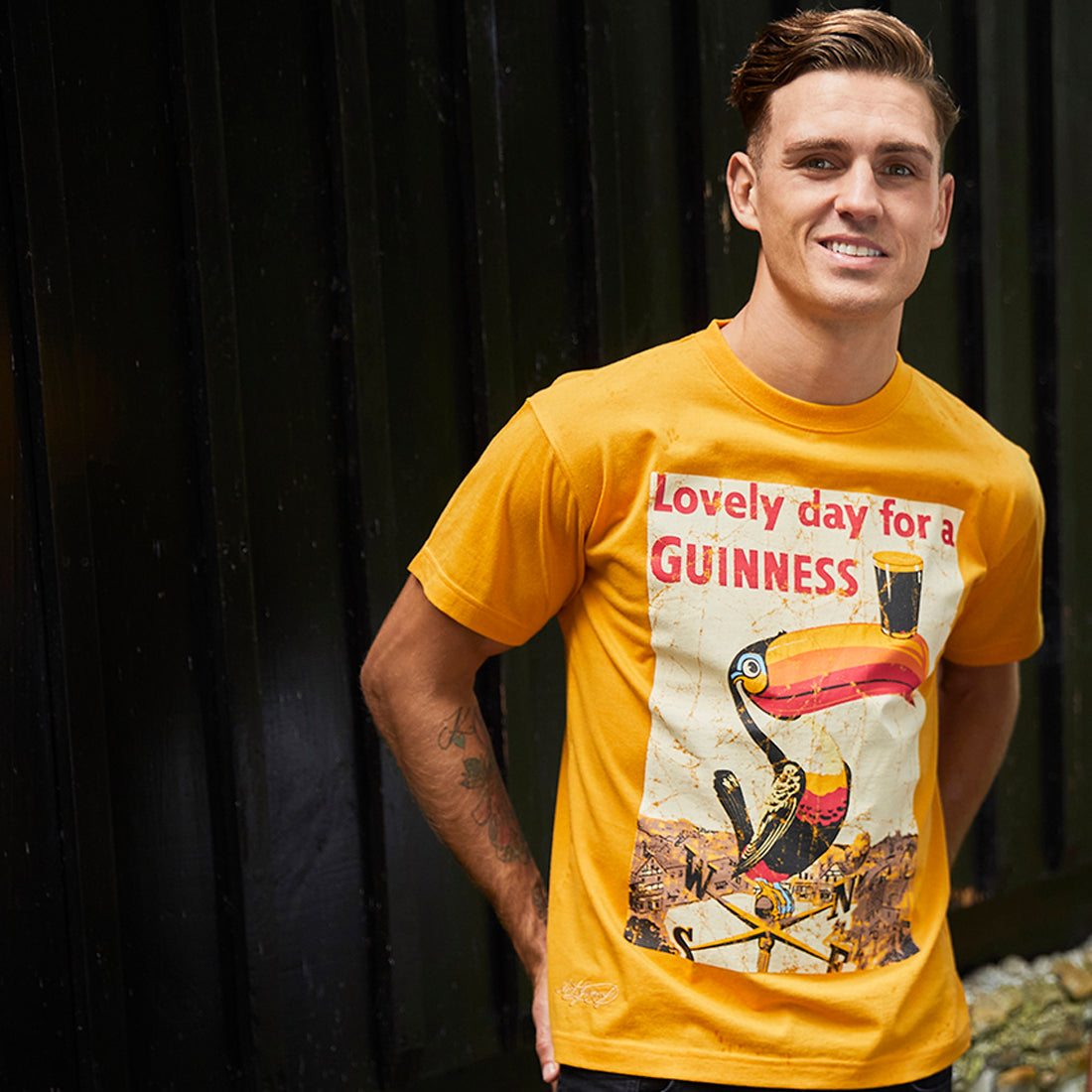 Man wearing the Guinness Lovely Day Toucan Golden T-Shirt front view, smiling against a dark background