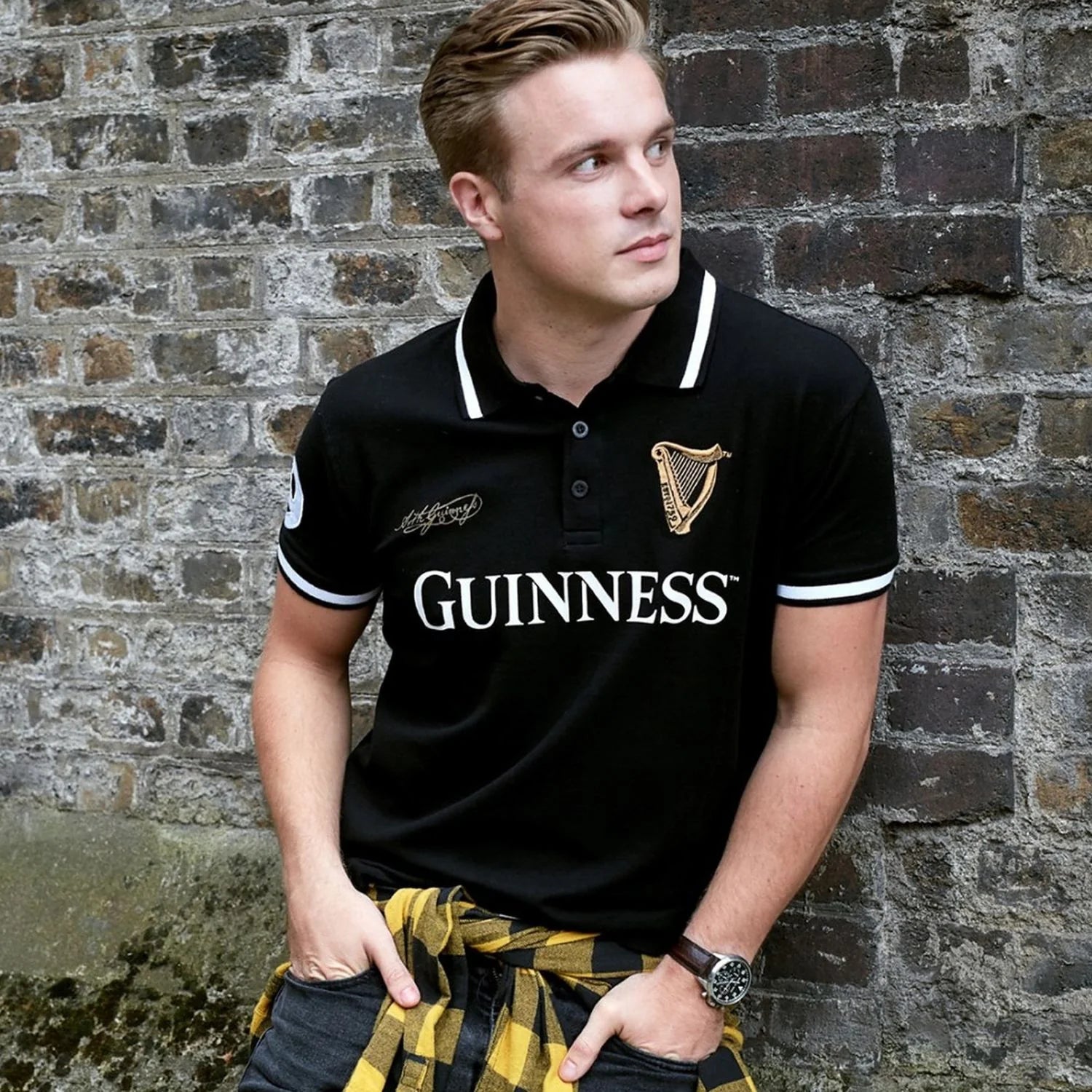 Male model wearing black Guinness polo shirt with white collar trim and crest emblem against a brick wall