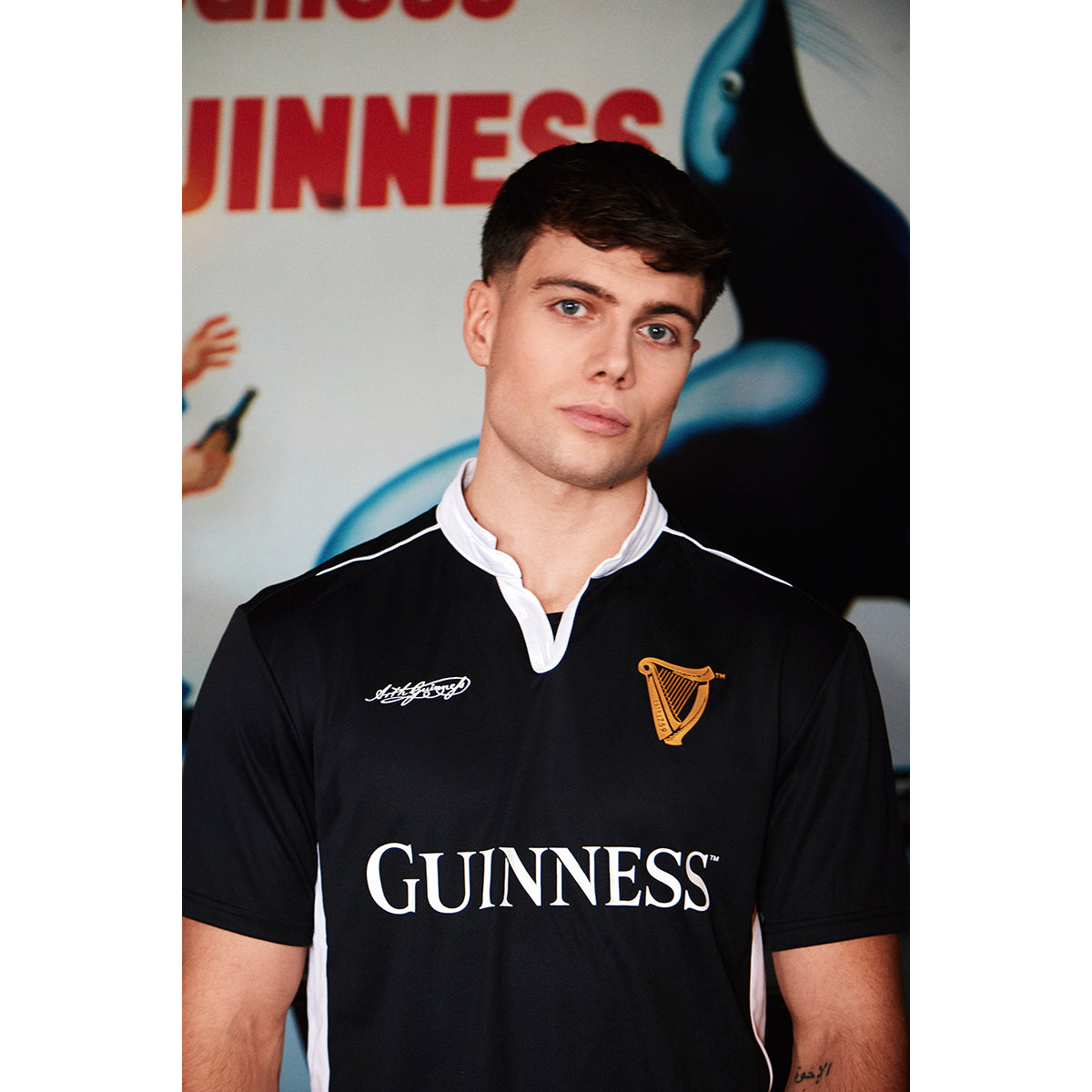Male model in black Guinness performance top with white collar trim and gold harp crest backdrop of Guinness branding.