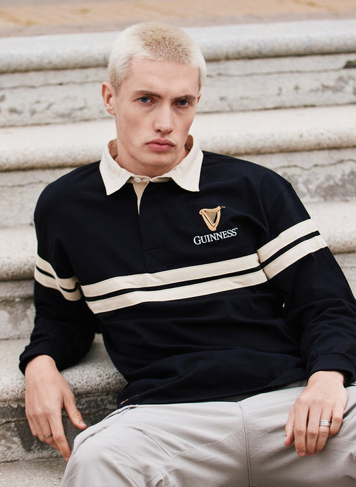 Model wearing Guinness Harp long sleeve rugby shirt in black with cream stripes and beige collar, seated on concrete steps
