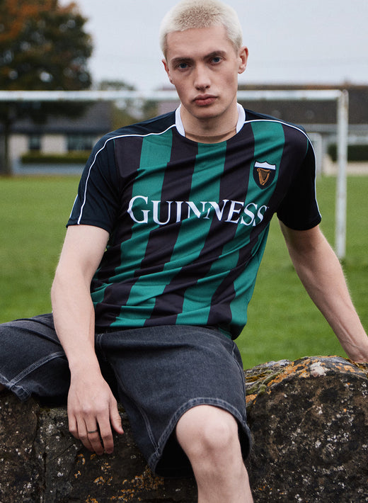 Man wearing Guinness Retro Striped Performance Top with green and black vertical stripes sits on a stone wall outdoors.