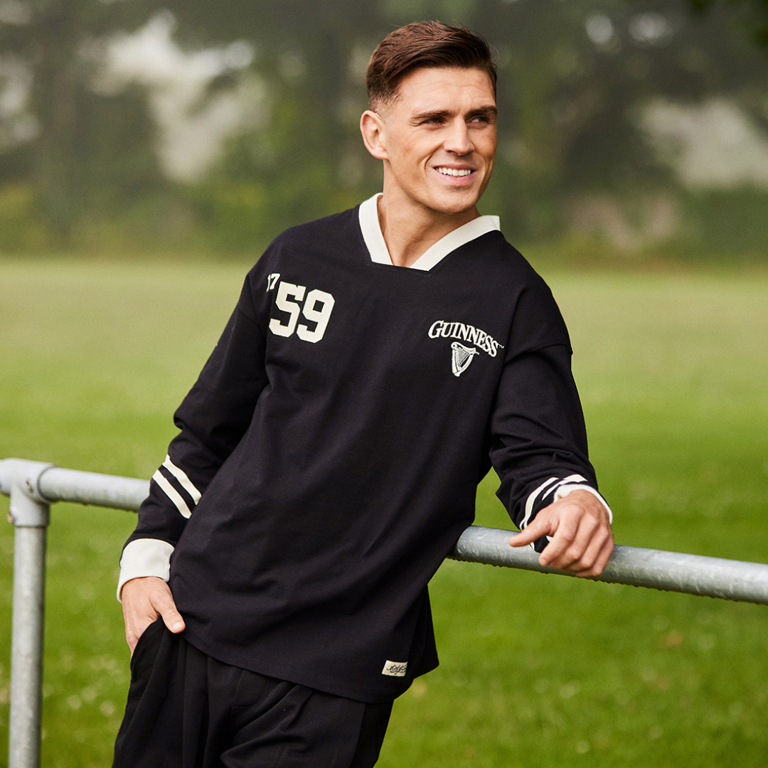 Smiling man wearing a black Guinness Toucan football top with white trim posing by a fence.