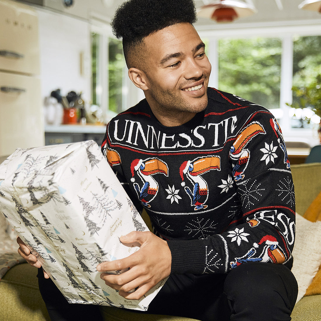 man with black hair sitting on a green couch and looking to the left in a room holding a present wrapped in white and  silver wrapping paper wearing a black sweater with toucans wearing red Christmas hats and Guinness time written in white