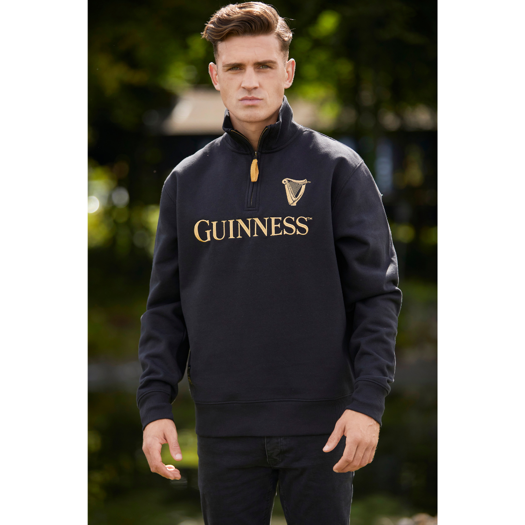 Male model standing outdoors in a black Guinness quarter zip sweatshirt featuring a gold harp crest and Guinness logo on the chest.