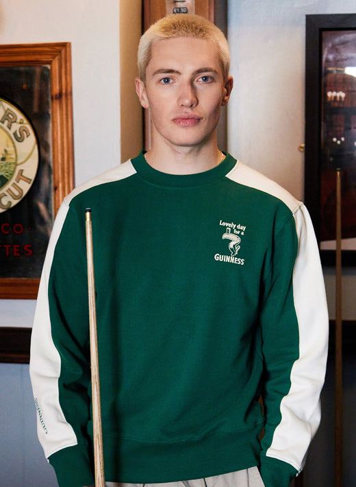 Front view of a man wearing a green and white Guinness Toucan sweatshirt with a chest logo, holding a pool cue.