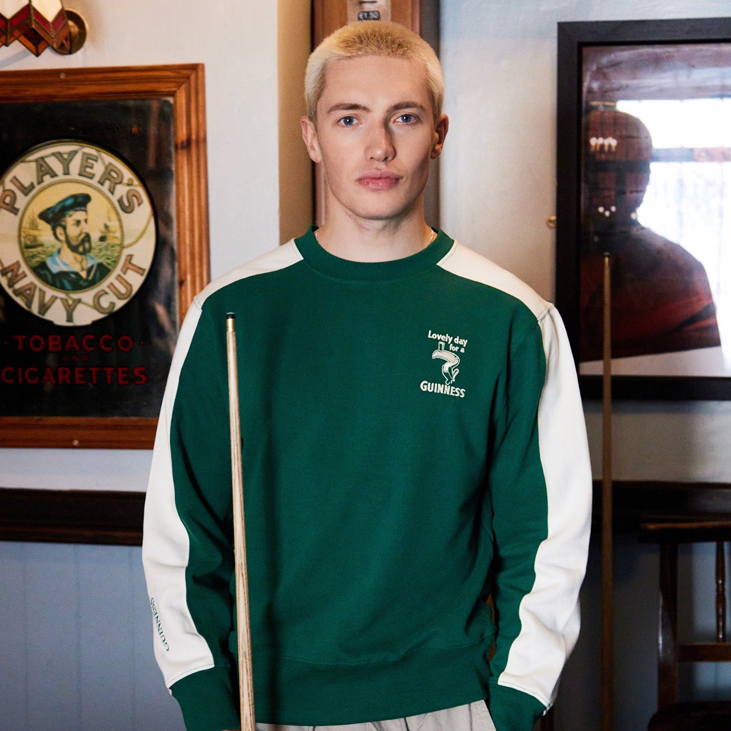 Front view of a man wearing a green and white Guinness Toucan sweatshirt with a chest logo, holding a pool cue.