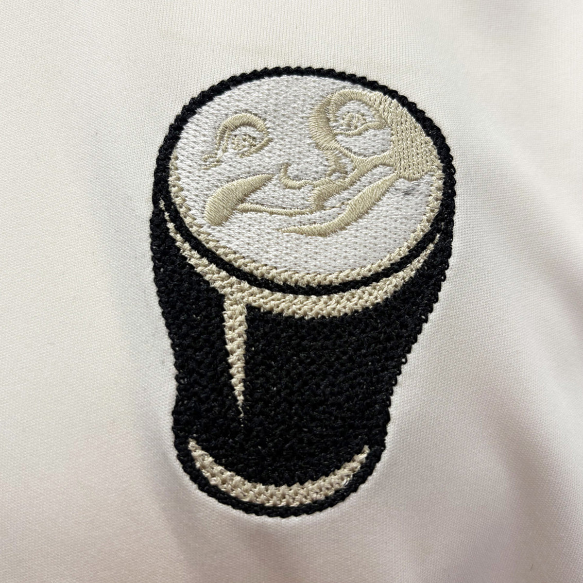 Close up of cream fabric showing an embroidered circular patch depicting a pint of beer.
