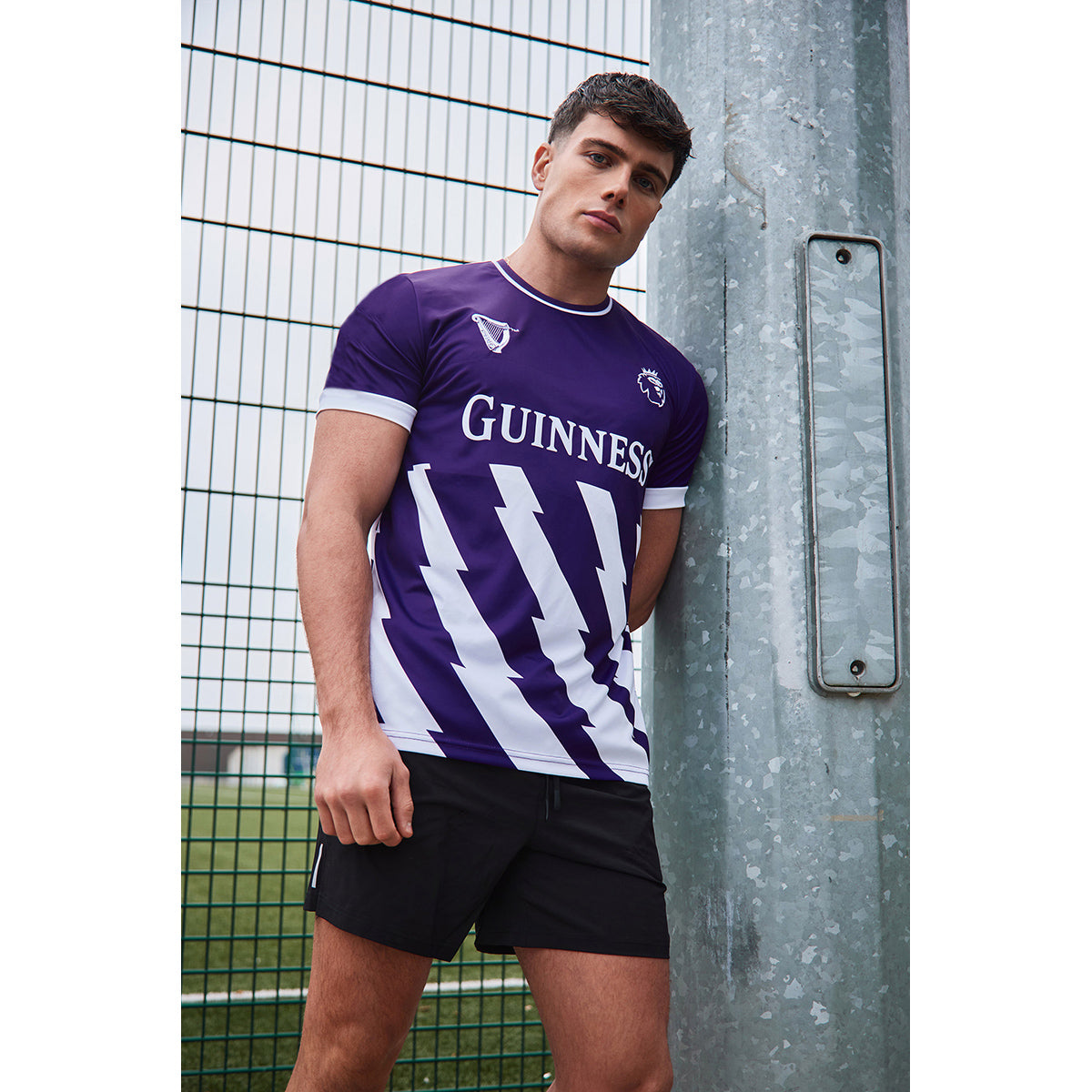 Male model wearing a purple Guinness football top with white diagonal stripes leaning against a metal post outdoors.