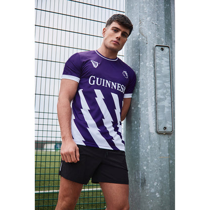 Male model wearing a purple Guinness football top with white diagonal stripes leaning against a metal post outdoors.