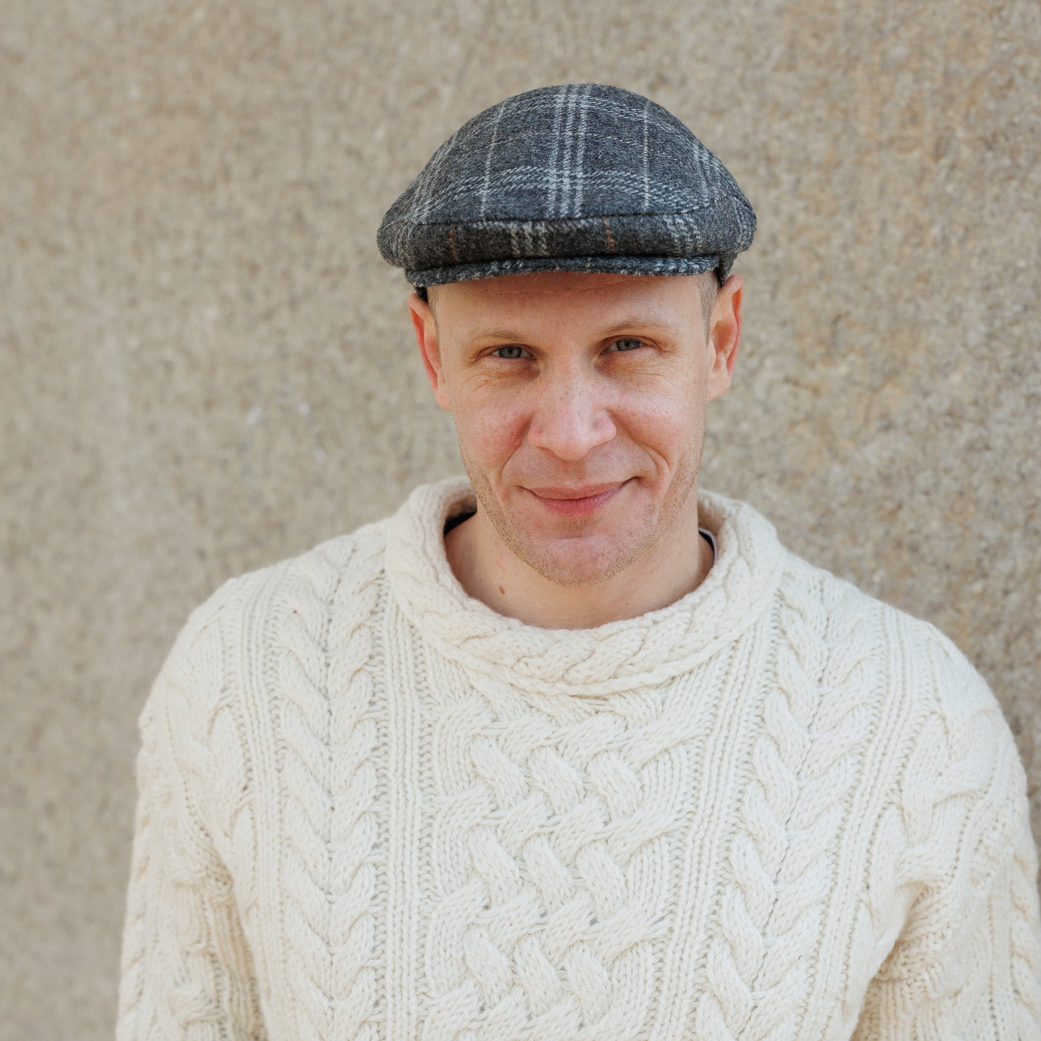Man wearing a gray flat cap and cream cable-knit sweater smiling