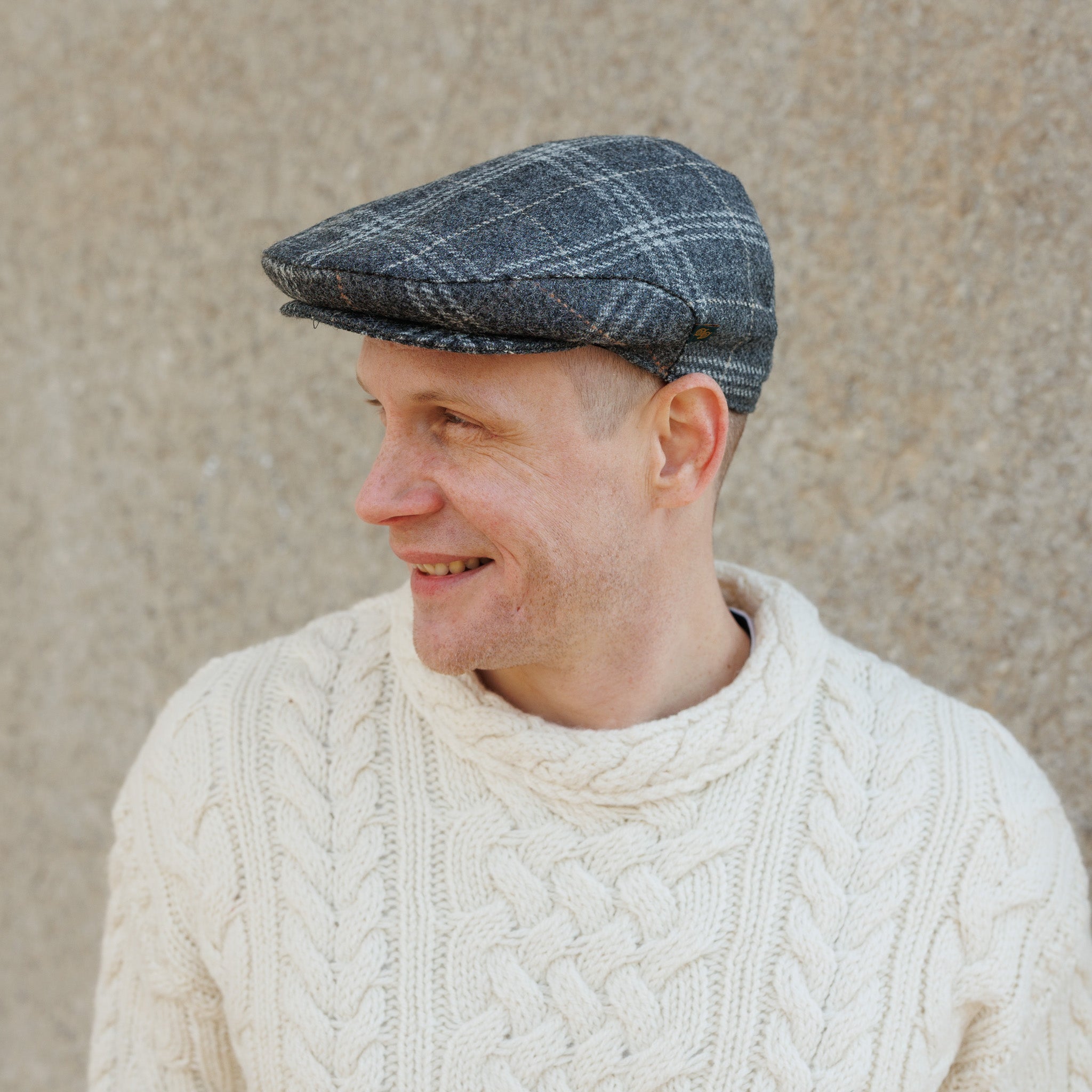 Man wearing a gray plaid flat cap and cream cable knit sweater smiling sideways