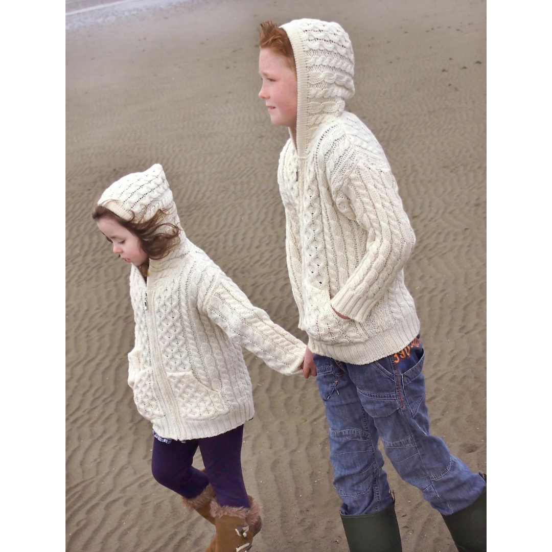 Two children in cream knit hoodies holding hands walking on sandy beach